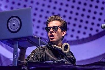 Mark Ronson onstage wearing sunglasses, infront of a purple background.