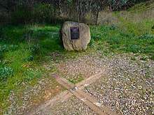 A boulder with a plaque is in the background, with a wooden cross embedded in the ground in the foreground.