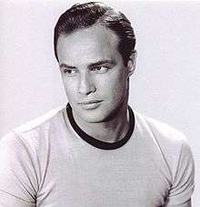 Black-and-white photo of Marlon Brando in 1953—a strong, handsome white man with dark eyes and dark hair, thick lips, square face and broad shoulders, wearing a light-colored shirt, around 28 years of age.