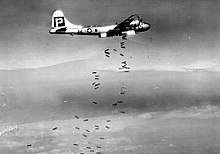 Black and white photograph of a World War II-era bomber releasing bombs. The bombs are falling in a scattered pattern.