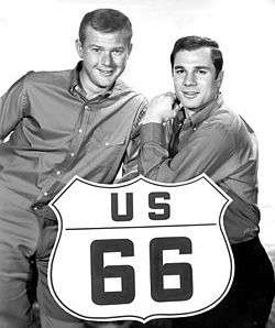 Black-and-white (greyscale) photo of blond-haired man at the left of the U.S. Route 66 large sign and a black-haired man at right
