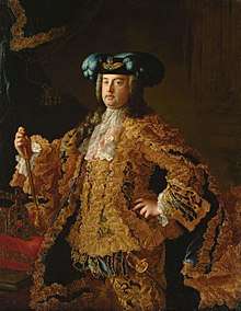 A man poses in gold laced Roman-German-Imperial coronation robes, the Crown of the Holy Roman Emperor lies on a cushion to his right.