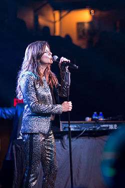 A woman with long brown hair wearing a silver jacket and pants, singing into a microphone