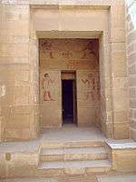 Rear wall of a vestibule in the tomb is visible through a doorway. On this wall, above and to the sides of a second door leading farther into the tomb, are murals painted in color. They show Khnumhotep and Niankhkhum seated at table above the second door, then they stand facing one another across the second door, each holding his staff of authority.