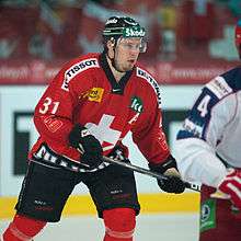 A Caucasian male ice hockey player shown from the knees up. He is wearing a red and white sweater with a dark helmet.
