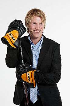 An ice hockey player standing directly in front of the camera while leaning on an ice hockey stick. He is wearing a black suit with a blue shirt and tie and is wearing black and orange hockey gloves.