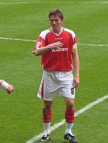 A brown-haired footballer in a red-and-white shirt, shorts and socks with his right arm across his chest, standing on a football pitch