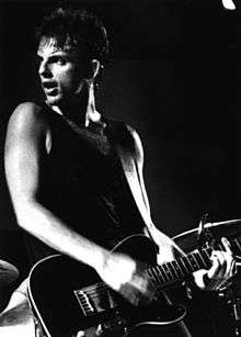 A 27-year-old man is shown in three-quarter shot. He is playing a guitar while leaning back slightly and his head is turned to his right. He wears a black singlet, strums the strings with a guitar pick in his right hand and holds strings at the fret board with his left. Behind him is part of a cymbal.