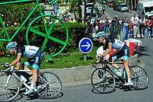 A pair of road racing cyclists in the team's black, white, and blue jersey, riding around a traffic island that features a large green sculpture of a bicycle. Spectators and motor vehicles are visible behind roadside barricades in the background.
