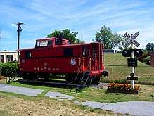 A red caboose on a short set of railroad tracks with a crossing signal at right. Behind them are a chainlink fence and a small rise