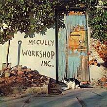 A colour photograph mid shot of a graffitied wall, a shovel leaning against it, and dilapidated blue door with a cat in front of it