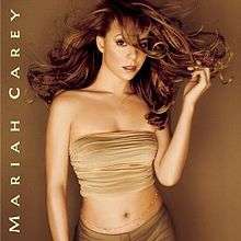 Image shows Carey standing in front of a brown/gold background in a beige sleeveless and mid-baring top, with darker matching pants. Her hair is long and golden-auburn, and is flowing in the air. her left hand is touching the flowing tips of her hair. She has a jeweled belt along her naval, with the words "Mariah Carey" written along the album cover (forgetting the album title).