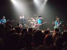 Slightly wonky image of a five-man band performing on-stage in front of rows of fans.