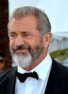 A photograph of Mel Gibson, sporting a beard and wearing a suit with matching bow tie while focused in another angle, smiling.