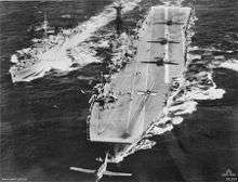 Aerial view of an aircraft carrier and a frigate sailing in close formation. Three propeller aircraft are lined up on the deck of the carrier, while a fourth has just been launched from the catapult.