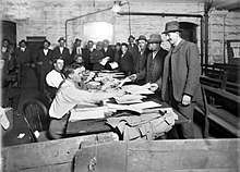 Men in suits and hats file past other men sitting at a long table, handing over paperwork.