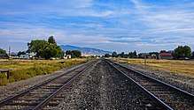 A pair of railroad tracks come from either side of the camera at the bottom corners to join into one midway us the image. On either side are two large grassy areas; on the right hand one a small wooden sign says "Melrose". There are streets with low buildings and some cars beyond the grassy areas, mountains in the distance, and blue sky with cirrus clouds above.