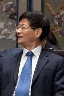a man with a wavy haircut, wearing glasses, a white shirt, a suit and a blue tie
