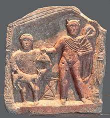 Crude stone relief carving of two standing male figures, facing the viewer and flanked by columns under a peaked roof; the taller figure is mostly nude and holds a bag or purse toward the shorter figure, who holds a goat by the horns