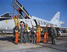 The astronauts pose in alphabetical order in front of a delta-winged white jet aircraft. They are holding their flight helmets under their arms. The three Navy aviators wear orange flight suits; the Air Force and Marine ones wear green.