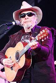 An old man in a cowboy hat, dark glasses and a dark jacket playing a guitar on stage