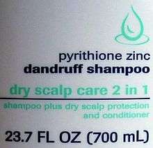 White slightly curved plastic with a green line drawing of a drop, the text "pyrithione zinc dandruff shampoo" in dark blue with "dry scalp care 2 in 1" in green, then a blue line. Underneath it is "shampoo plus dry scalp protection and conditioner" in smaller green type, and finally "23.7 FL OZ (700&nbsp;mL)" in larger blue type.