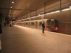 A train is waiting on a track in a subway station. Passengers are standing on the platform.
