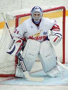 An ice hockey player standing partially crouched in goals. He is wearing a helmet, gloves and leg pads and a white uniform.