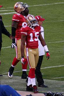 In the foreground, an American football player wearing a gold helmet, red jersey (number 15), gold pants and red socks walks along the sideline. Behind him, a man wearing a red cap and white T-shirt and another player in the same football uniform as the player in the foreground, walk in the same direction.
