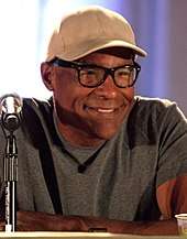 A man in a baseball cap, glasses and a grey T-shirt.