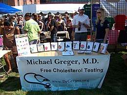 Photograph of Michael Greger at the D.C. Vegfest in 2007, standing in a white shirt in front of a booth whose banner states "Michael Greger, M.D." with "Free Cholesterol Testing" below it and his website at the bottom