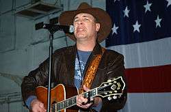 A middle-aged man in a cowboy hat and a leather jacket, playing a guitar and singing into a microphone in front of the American flag