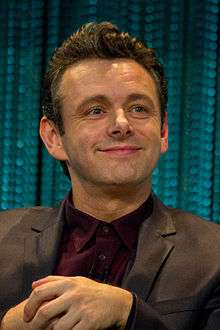 Michael Sheen, a caucasian male in his mid-40s with dark hair, wears a black suit and burgundy shirt.