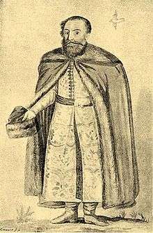 An elderly bearded man with a hat in his hand wearing a coat decorated with furs
