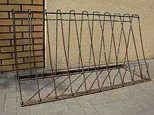A rusty metal A-frame with horizontal bars at the top and bottom joined by eleven vertical bars. It stands free on concrete in front of a join between a brick wall and a plaster wall.