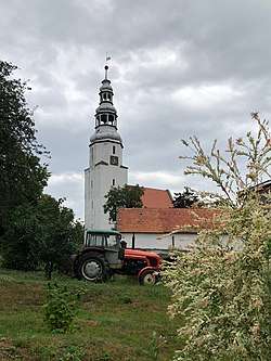 The tall white tower of the church in Mierczyce village having a clock face and standing near the red roof of the church as seen across a nearby yard where a tractor being parked in.