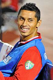 A young Latino man wearing a blue sleeveless T-shirt over a red long-sleeved T-shirt looks to his left and smiles.
