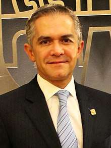 Miguel Ángel Mancera stands in front of a golden shield depicting an old Mexico City's shield. He looks directly to the camera and wears a black suit.