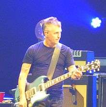 A male electric guitar player, Mike McCready, onstage with an electric guitar plugged into a guitar amplifier.
