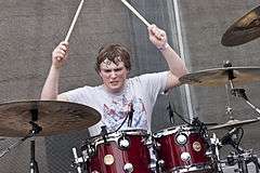 Mike Byrne—a Caucasian male 20 years old with short brunette hair with a grey T-shirt—drums behind a red drum kit.
