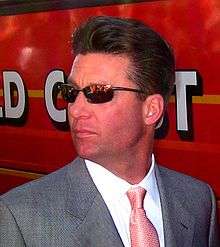 A man wearing sunglasses and a gray suit with a patterned red tie looking over his right shoulder