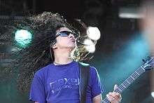 A male bass player, Mike Inez, performing at a concert. He has long curly hair and wears sunglasses.