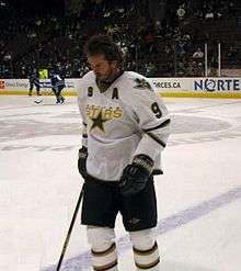 An ice hockey player stands on the ice. He is wearing a white jersey with the word "Stars" on the front in gold.
