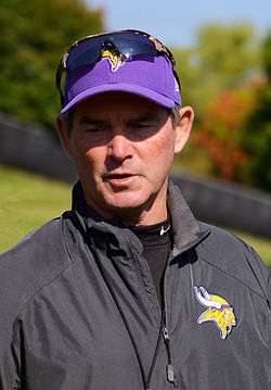 Color full-length photograph of Zimmer, gesturing with his hands, walking on a football field, wearing a grey pullover, grey shorts and purple baseball cap with the Minnesota Vikings logo.