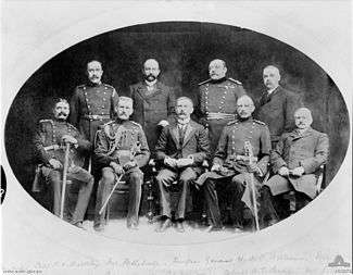 Formal group portrait of nine men, four sitting at the front and five standing behind. Three are wearing suits; the others are wearing formal double breasted military uniforms with sashes.
