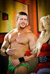A dark-haired, caucasian man with stubble is arm-in-arm with a blonde-haired woman who is wearing a dress in the style of Santa Claus. The man is wearing short green wrestling trunks, and is gesticulating towards in the woman in an apparent conversation.