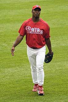 A dark-skinned man wearing a red baseball jersey and cap and white pinstriped baseball pants walking on a grass field