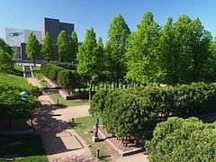 Hedge-lined paths with statues and other sculptures beside them run through much of the foreground of the image. In the background stands a gray metal art museum. The day is sunny and clear.