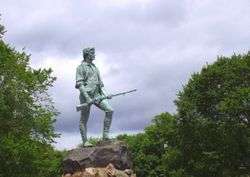 A weathered green statue is mounted on a rough stone pedestal in front of some trees. The statue is of a man holding a long gun.
