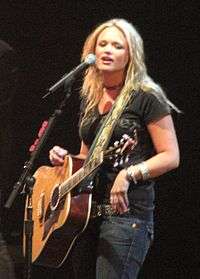 A woman with long blonde hair, wearing a black t-shirt and blue jeans, playing a guitar and singing into a microphone